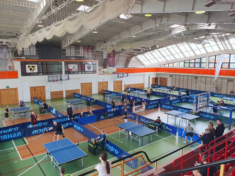 zdjęcie przedstawiające wiele stołów do tenisa stołowego rozstawionych w hali sportowej i zawodników biorących udział w turnieju